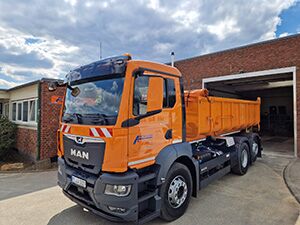Dreiseitenkipper - Apken - GmbH Ein Containerfahrzeug in orange steht vor einer Garage.