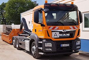 Nachläufer - Apken GmbH Ein Containerfahrzeug in orange steht mit heruntergelassenem Container vor einem Gebäude.